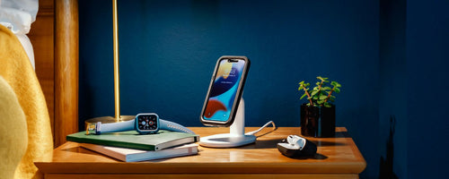 iPhone charging on a MagSafe wireless charging stand with Apple Watch and AirPods laying on side table