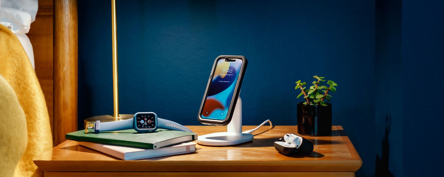 iPhone charging on a MagSafe wireless charging stand with Apple Watch and AirPods laying on side table