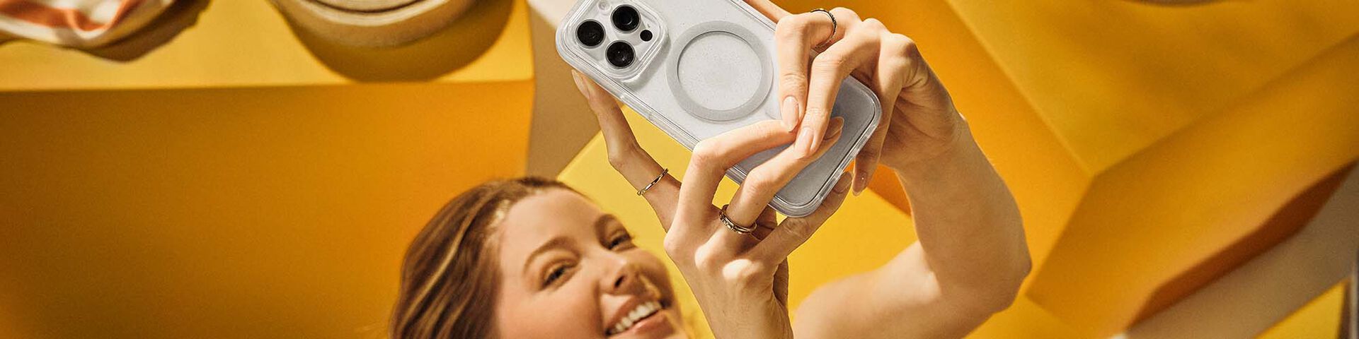 Woman taking a selfie on white iPhone 16 Pro Max in clear MagSafe OtterBox phone case on a yellow backgroung