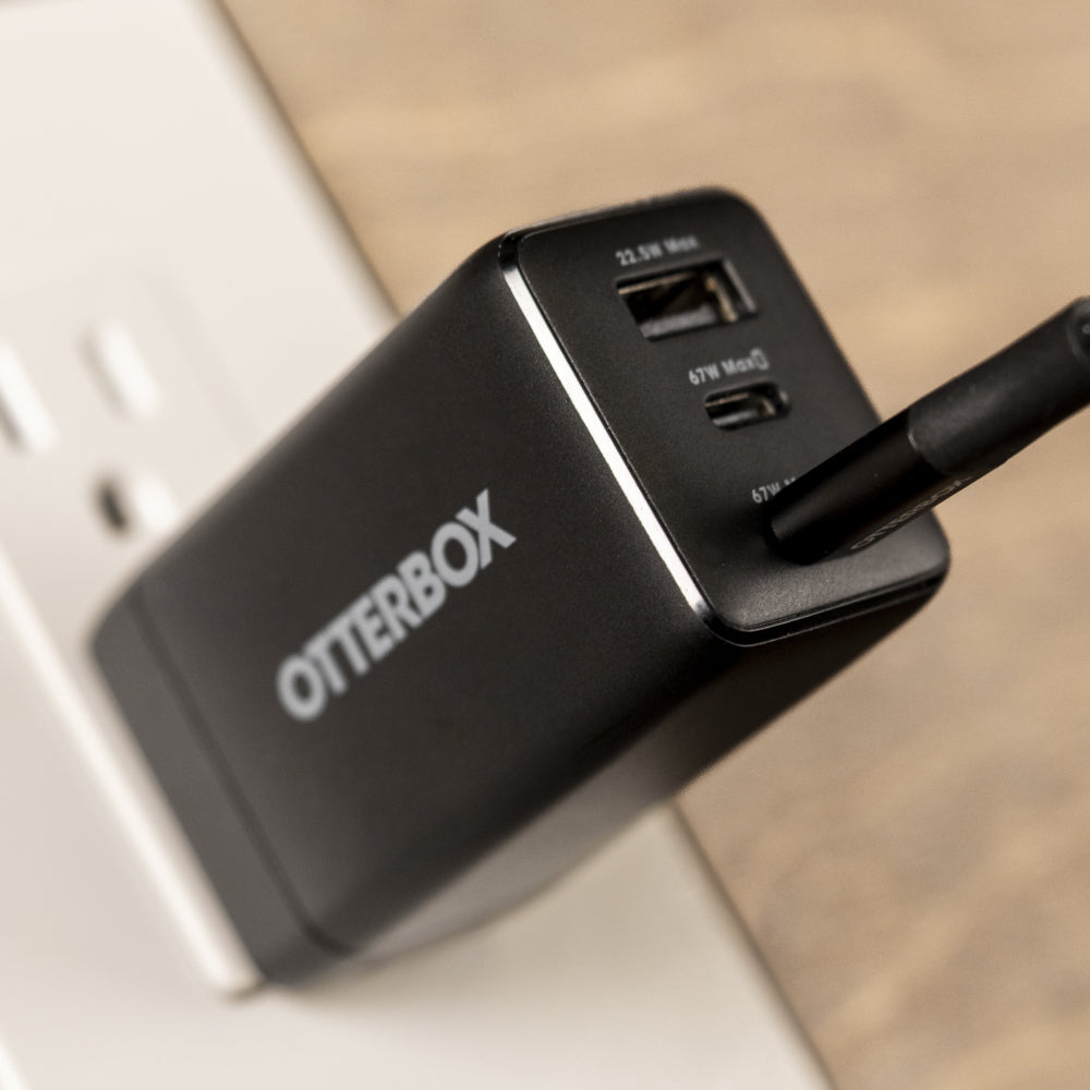 A close-up of a black Otterbox USB charger plugged into a white wall outlet, with a wooden surface blurred in the background.