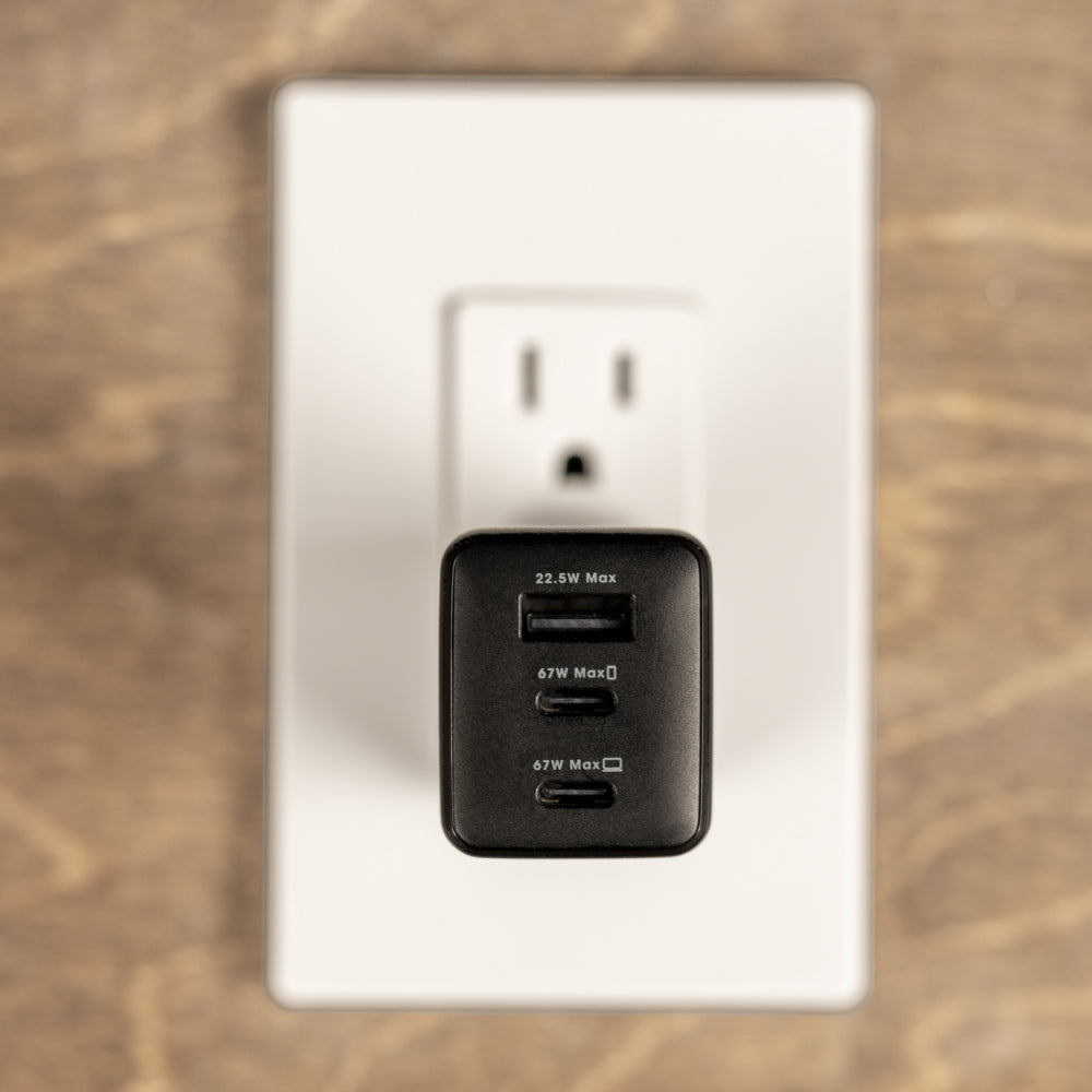 A black dual-port USB charger plugged into a white wall outlet on a wooden surface.