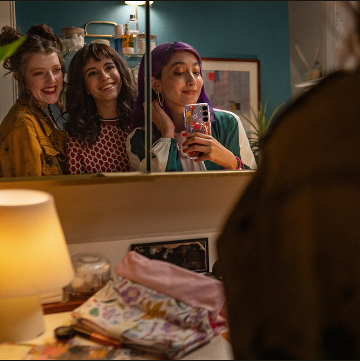 Three women taking a mirror selfie in a room with a lamp and decorative items.