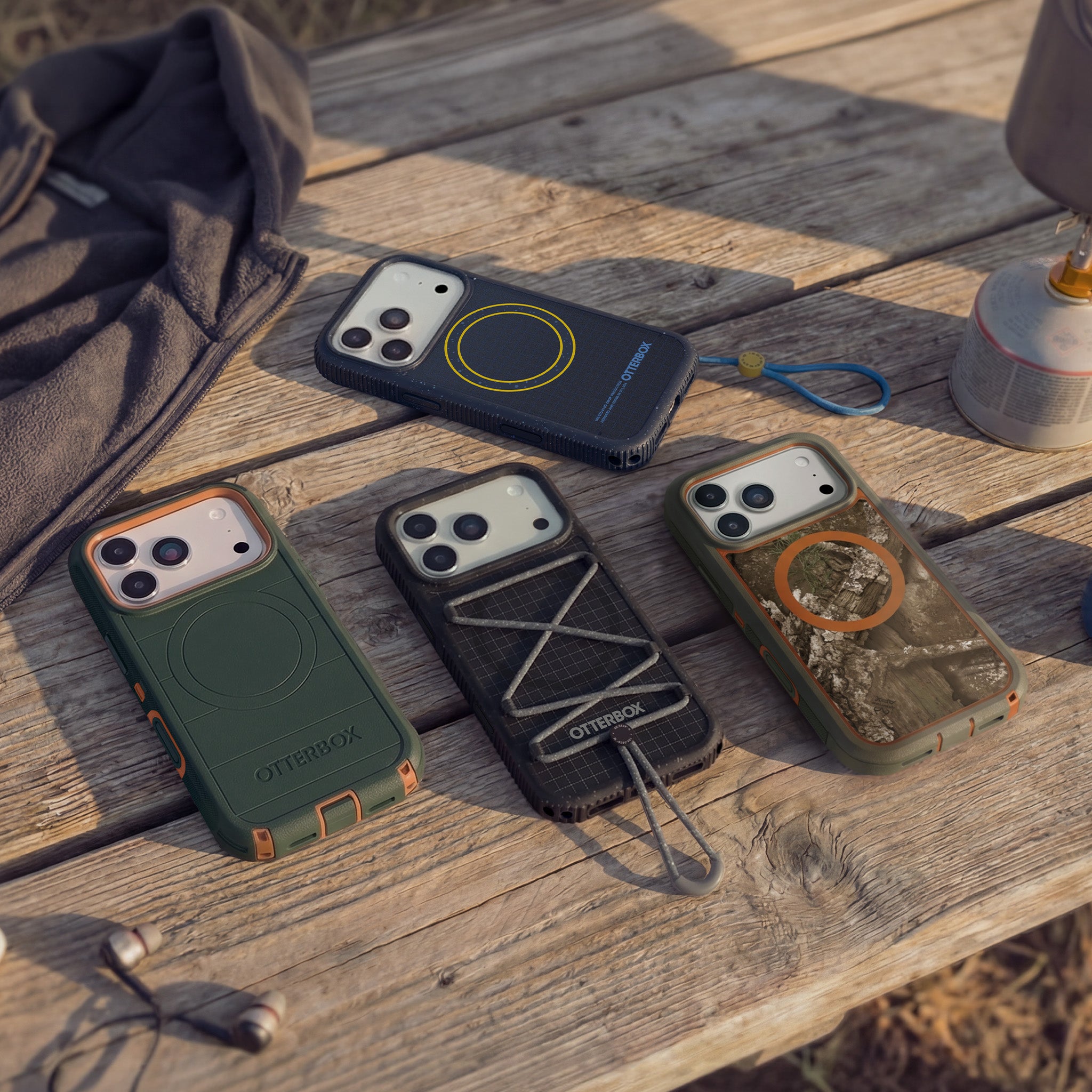 Four OtterBox iPhone cases in different color and material designs, arranged on a rustic wooden table outdoors with warm natural sunlight.
