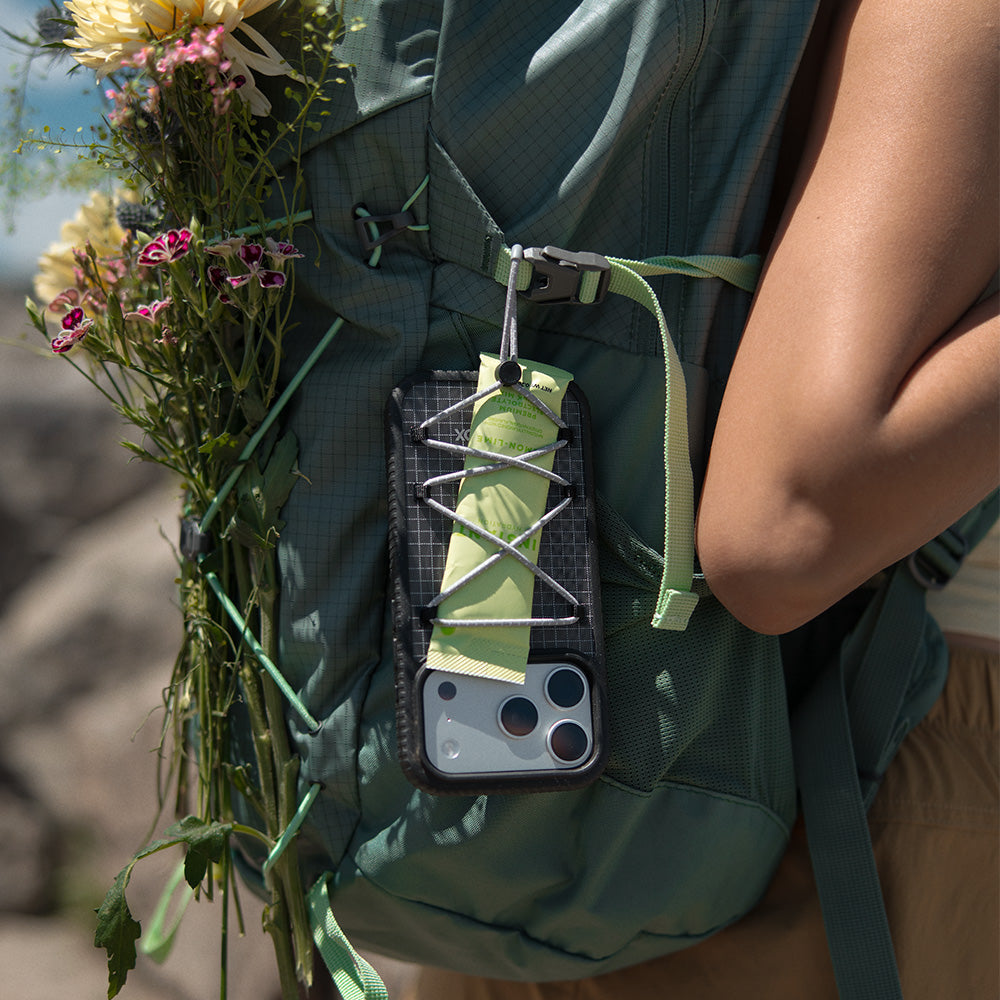 A close-up outdoor shot of a teal hiking backpack with a side-mounted phone pouch holding a smartphone and a light green loop. A person wearing the backpack is partially visible, along with a bouquet of flowers tucked into the pack.