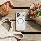 White iPhone in a clear protective case on a wooden picnic table beside a canvas tote bag, loaf of bread, and bouquet of flowers.