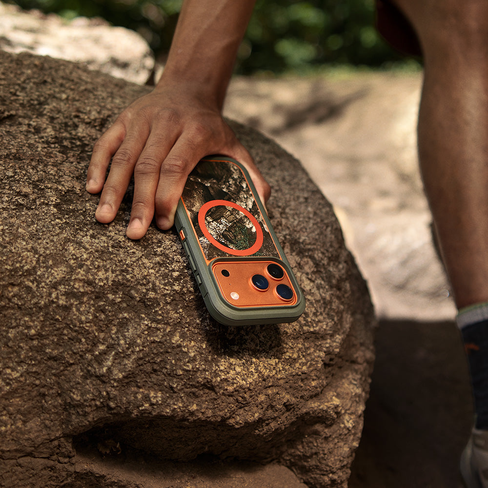 A rugged smartphone case with a camouflage pattern and orange accents rests on a large rock outdoors, with a person's hand and legs visible in the scene.
