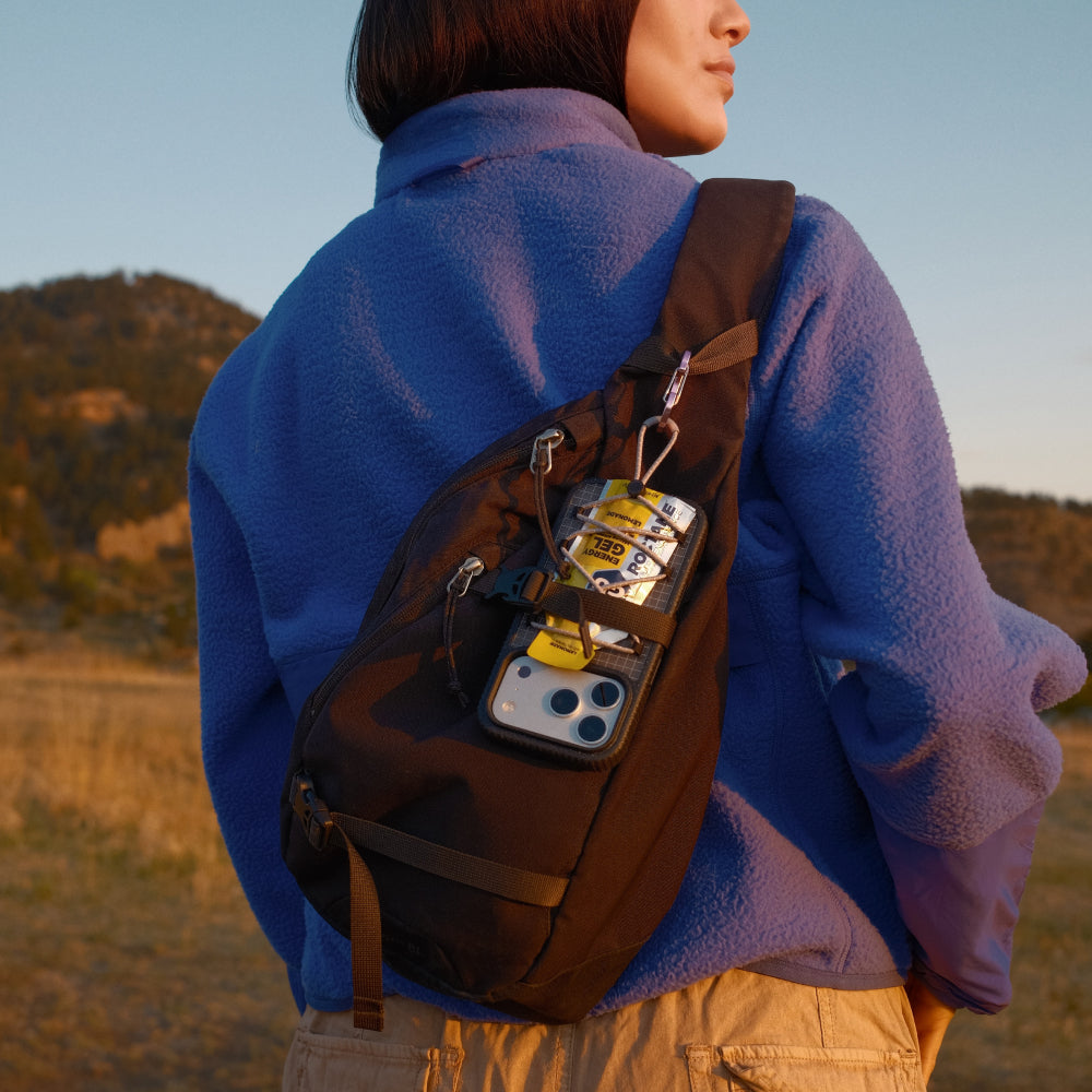 A person wearing a blue fleece jacket stands outdoors in a field at sunset, shown from the back while wearing a dark brown sling backpack with a phone pouch containing a phone and small accessories.
